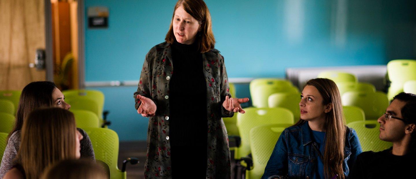 Students sitting at desks look towards a woman who stands speaking